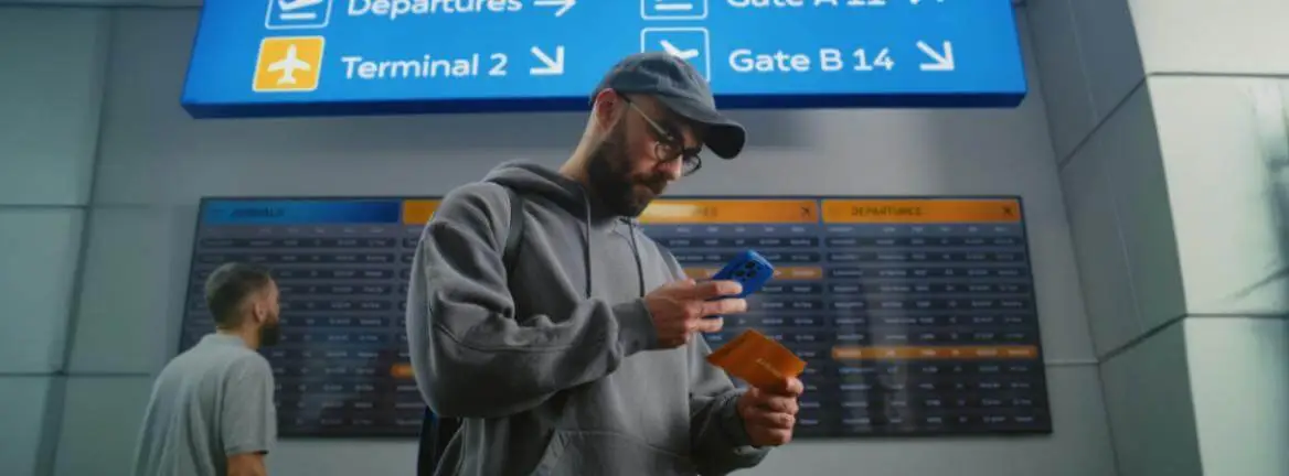 Passageiro no aeroporto usando app para acompanhar voo em tempo real, com painéis de voos ao fundo, transmitindo controle e atenção durante a viagem.